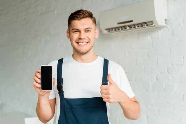 Instalador alegre que muestra el pulgar hacia arriba mientras sostiene el teléfono inteligente con pantalla en blanco - foto de stock