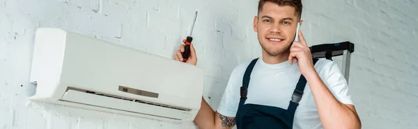 Colpo panoramico di felice installatore tenendo cacciavite mentre si parla su smartphone vicino condizionatore d'aria — Foto stock