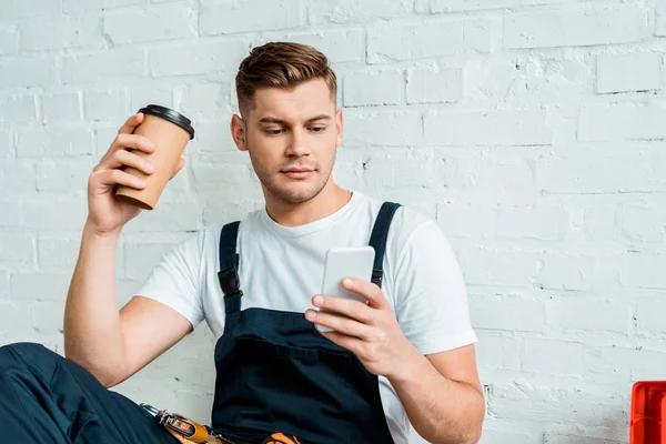Schöner Installateur schaut aufs Smartphone, während er Pappbecher in der Hand hält — Stockfoto