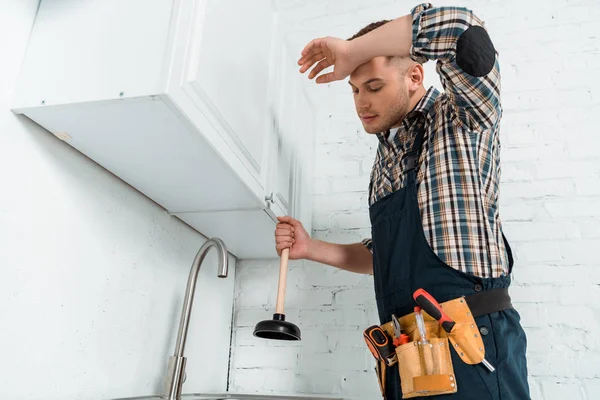 Уставший монтажник держит плунжер рядом с краном на кухне — стоковое фото