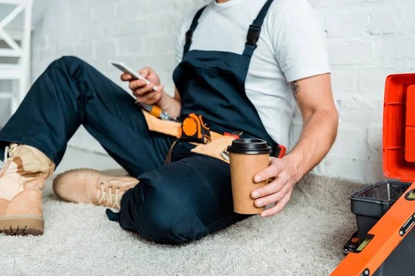 Ausgeschnittene Ansicht eines Installateurs, der auf Teppich sitzt, während er Pappbecher hält und Smartphone benutzt — Stockfoto