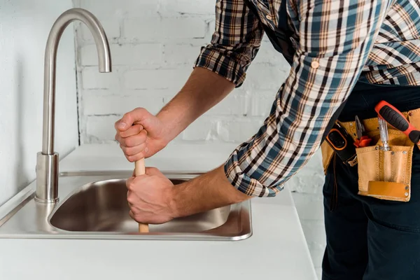 Vista recortada del plomero sosteniendo el émbolo cerca del grifo y el fregadero en la cocina - foto de stock