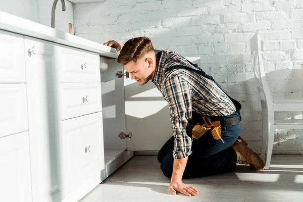 Tired installer standing on knees while working near kitchen cabinet — Stock Photo