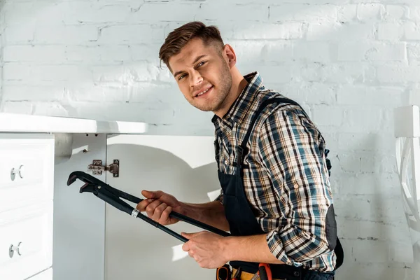 Glücklicher Installateur, der eine Gleitgelenkzange hält, während er in der Nähe von Küchenschränken arbeitet — Stockfoto