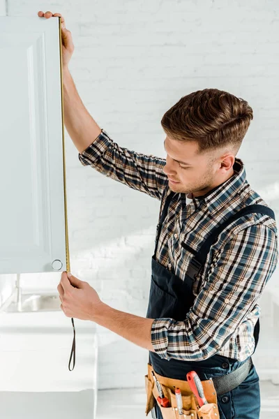 Beau installateur en salopettes mesurant blanc armoire de cuisine porte — Photo de stock