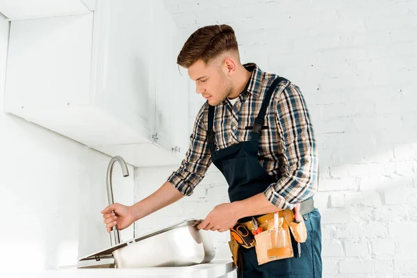 Instalador bonito segurando pia metálica perto da torneira na cozinha — Fotografia de Stock