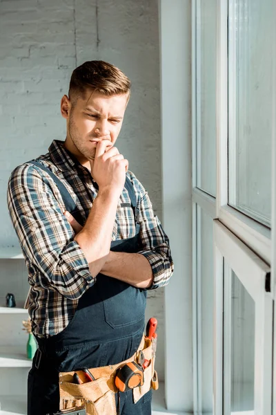 Nachdenklicher Installateur berührt Gesicht, während er in der Nähe von Fenstern denkt — Stockfoto