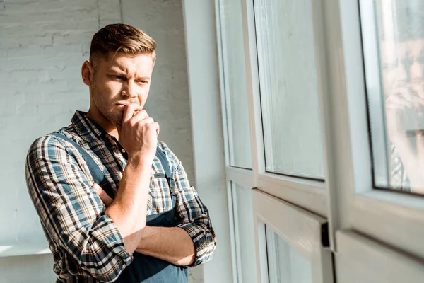 Selektiver Fokus des nachdenklichen Installateurs, der das Gesicht berührt, während er in der Nähe von Fenstern denkt — Stockfoto