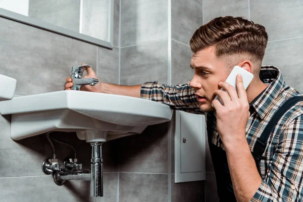 Bonito instalador tocando torneira e falando no smartphone — Fotografia de Stock