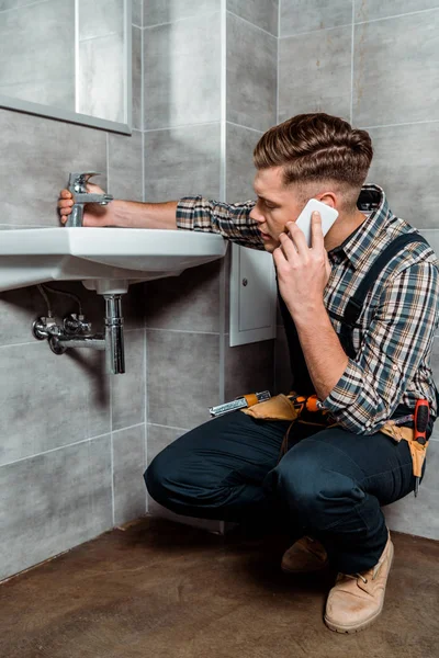 Bello installatore seduto vicino al lavandino e toccare rubinetto mentre si parla su smartphone — Foto stock