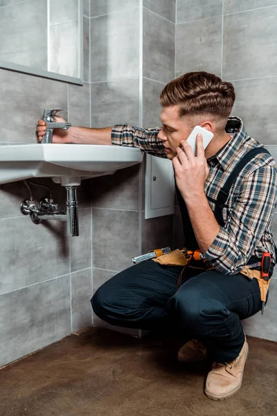 Instalador guapo sentado cerca del fregadero y tocar el grifo mientras habla en el teléfono inteligente - foto de stock