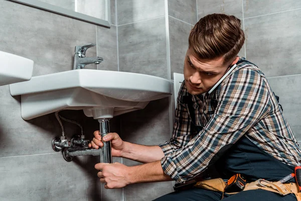 Installer touching pipe while talking on smartphone in bathroom — Stock Photo