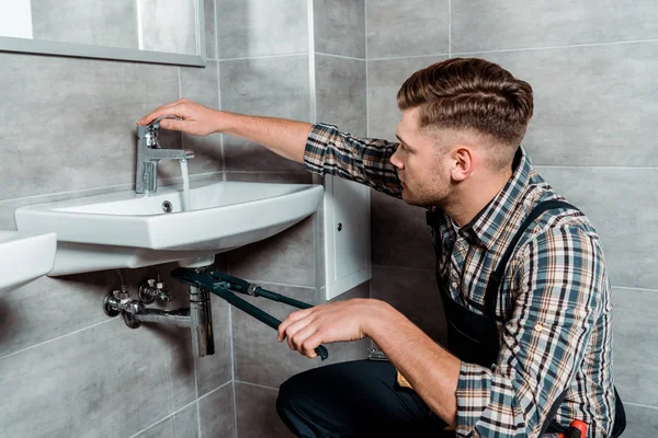 Seitenansicht des Installateurs, der eine Gleitfugenzange in Rohrnähe hält und den Wasserhahn im Badezimmer berührt — Stockfoto