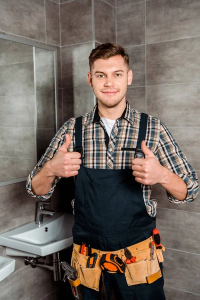 Installateur heureux avec ceinture d'outils debout dans la salle de bain et montrant pouces vers le haut — Photo de stock