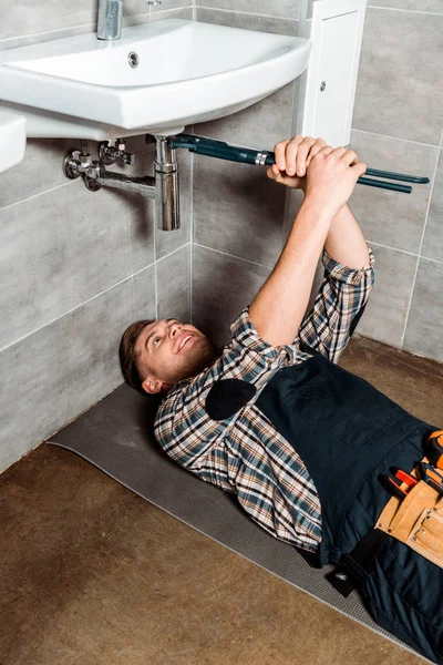 Glücklicher Installateur hält Gleitgelenkzange in Rohrnähe, während er im Badezimmer auf dem Boden liegt — Stockfoto