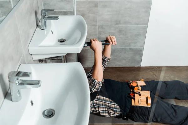 Обрезанный вид монтажника, держащего скользящие шарнирные плоскогубцы возле трубы, лежащей на полу в ванной комнате — стоковое фото