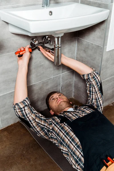 Instalador guapo sujeción alicates mientras está acostado en el suelo en el baño - foto de stock