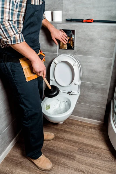 Обрезанный вид сантехника держа вантуз при нажатии кнопки смыва рядом с туалетом — стоковое фото