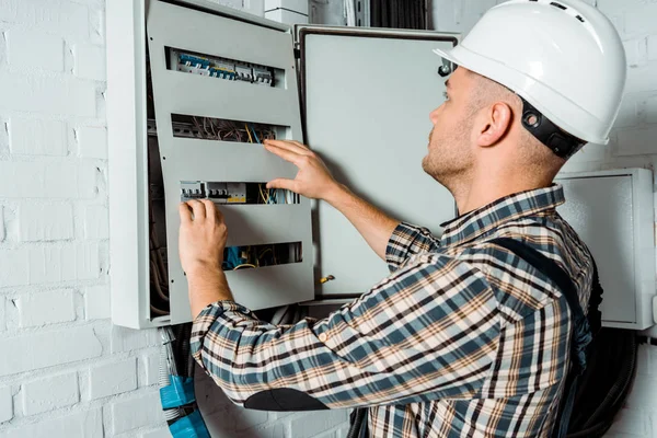 Electrician in safety helmet looking at switchboard near brick wall — Stock Photo