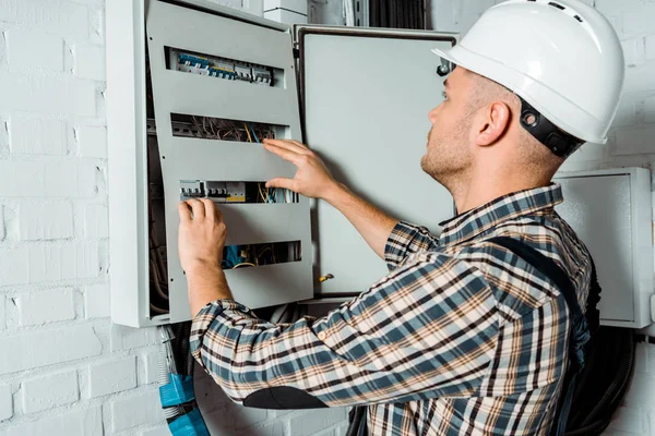 Electricista en casco de seguridad mirando la centralita cerca de la pared de ladrillo - foto de stock