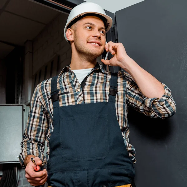 Fröhlicher Installateur im Schutzhelm hält Schraubenzieher während er mit dem Smartphone spricht — Stock Photo