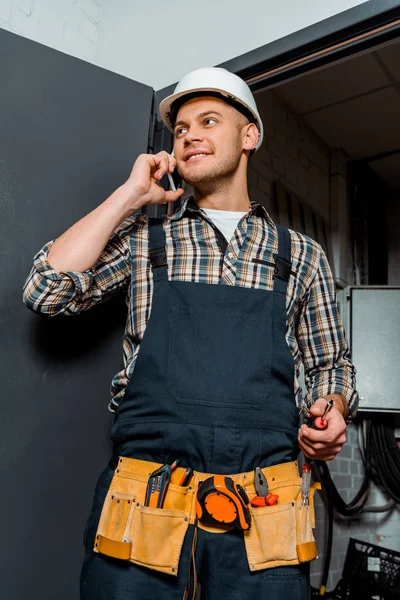 Happy installer in safety helmet holding screwdriver while talking on smartphone — Stock Photo