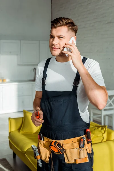 Sol en el instalador guapo en overoles con cortador y hablando en el teléfono inteligente - foto de stock