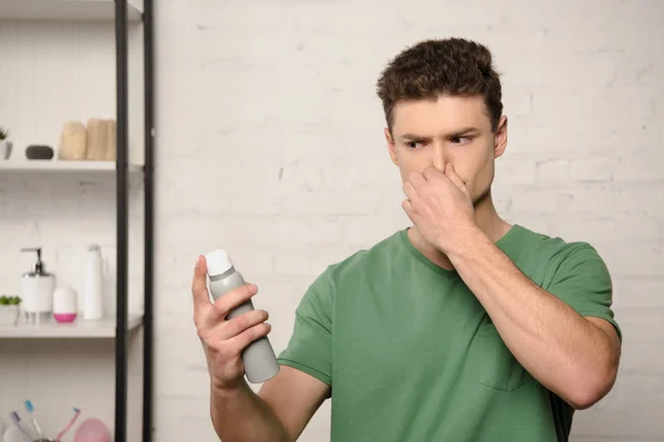 Unzufriedener junger Mann stopft Nase mit der Hand, während er Deo hält — Stockfoto