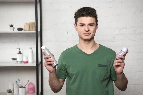 Positiver junger Mann im grünen T-Shirt, der in die Kamera schaut, während er Deos in der Hand hält — Stockfoto