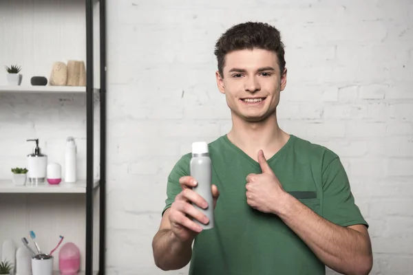 Sorridente giovane uomo mostrando pollice in su, mentre tenendo deodorante e guardando la fotocamera — Foto stock
