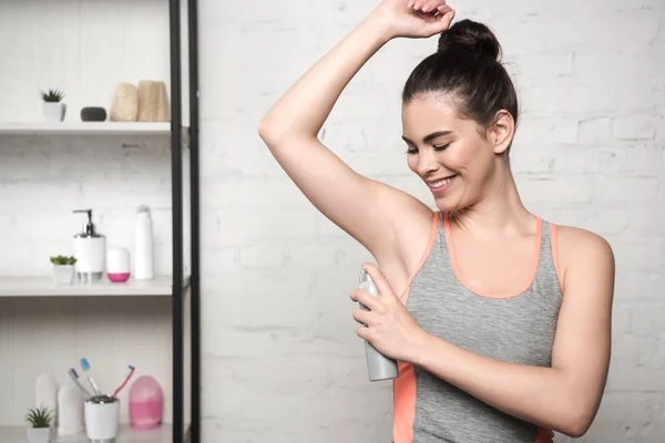 Donna sorridente in camicia grigia senza maniche spruzzando deodorante su ascelle — Foto stock