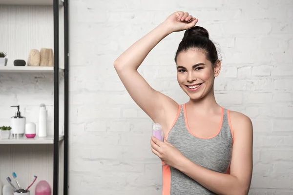 Happy woman smiling at camera while applying deodorant on underarm — Stock Photo