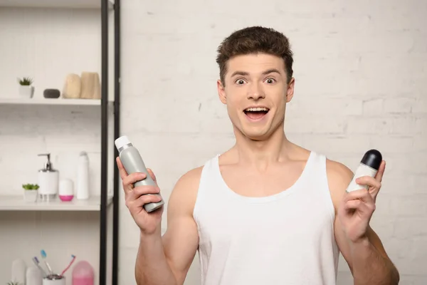 Excited young man in white sleeveless shirt looking at camera while holding deodorants — Stock Photo