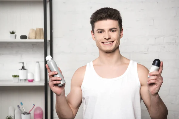 Young man in white sleeveless shirt holding deodorants and smiling at camera — Stock Photo