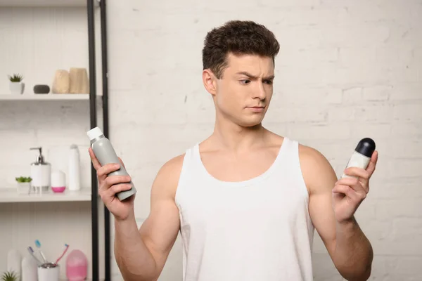 Skeptical young man in white sleeveless shirt holding deodorants — Stock Photo