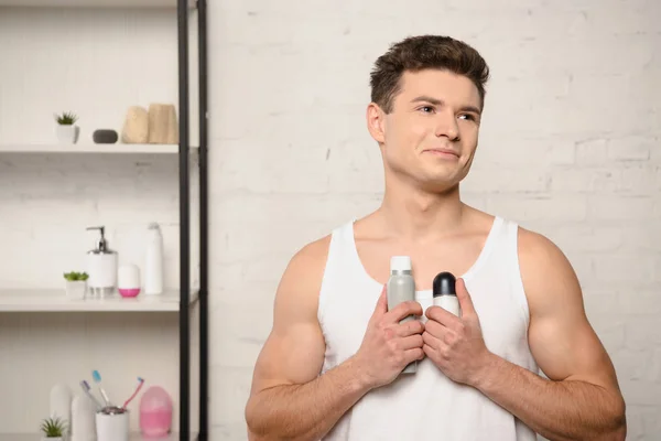 Giovane uomo sorridente in camicia bianca senza maniche guardando altrove mentre tiene in mano i deodoranti — Foto stock