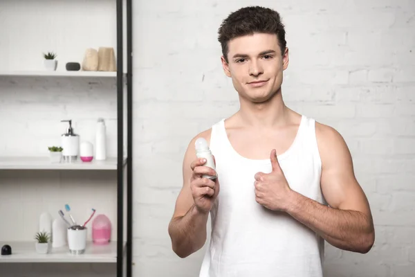 Positive man in white sleeveless shirt showing thumb up while holding deodorant — Stock Photo
