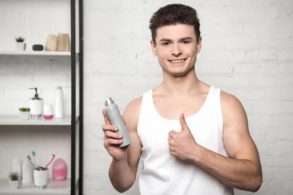 Hombre sonriente con camisa blanca sin mangas mostrando el pulgar hacia arriba mientras sostiene el desodorante - foto de stock