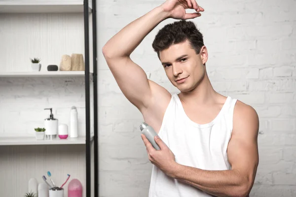 Young man in white sleeveless shirt looking at camera while spraying deodorant on underarm — Stock Photo