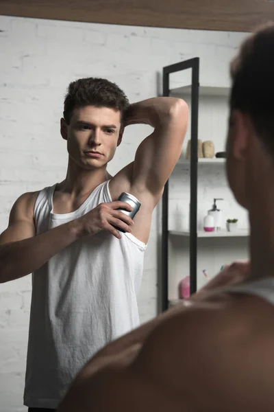 Young man in white sleeveless shirt applying deodorant on underarm while looking at mirror — Stock Photo