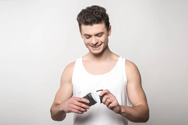 Hombre sonriente con camisa blanca sin mangas abriendo desodorante aislado en gris - foto de stock