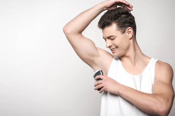 Uomo sorridente in camicia bianca senza maniche che applica deodorante su ascelle isolate su grigio — Foto stock