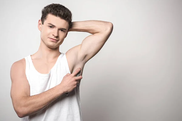 Handsome man in white sleeveless shirt applying deodorant on underarm and smiling at camera on grey background — Stock Photo