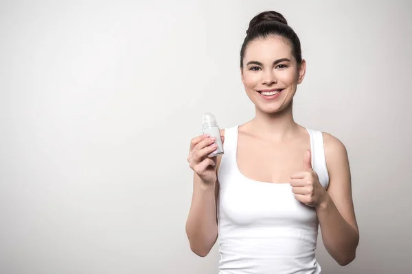 Mujer sonriente mirando a la cámara mientras sostiene el desodorante y muestra el pulgar hacia arriba aislado en gris - foto de stock