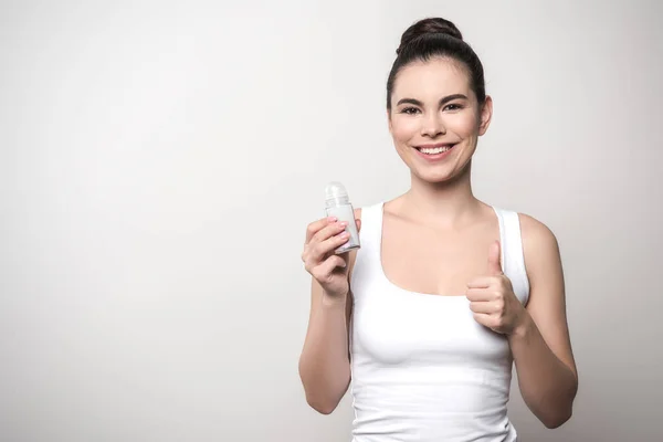 Lächelnde Frau blickt in die Kamera, während sie Deo in der Hand hält und den Daumen vereinzelt auf grau zeigt — Stockfoto