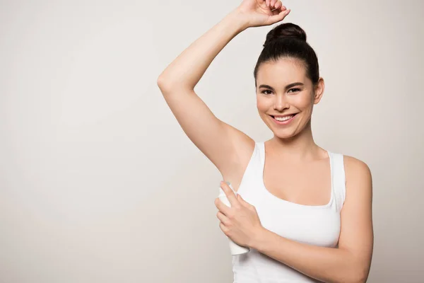 Cheerful woman smiling at camera while applying deodorant on underarm isolated on grey — Stock Photo