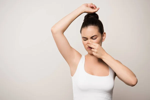 Displeased woman plugging nose with hand while looking at underarm isolated on grey — Stock Photo