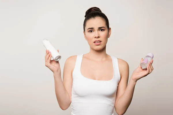 Donna infelice guardando la fotocamera mentre tiene i deodoranti isolati sul grigio — Foto stock