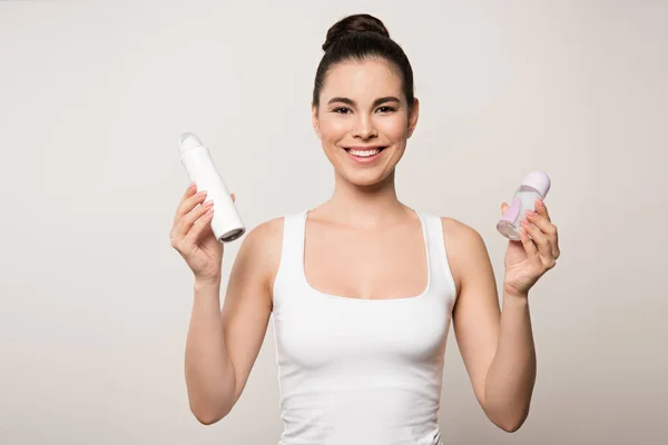 Радостная женщина улыбается в камеру, держа в руках дезодоранты, изолированные на сером — стоковое фото