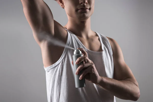 Teilbild eines jungen Mannes im weißen ärmellosen Hemd, der Deo auf den Achselarm sprüht, isoliert auf grau — Stockfoto